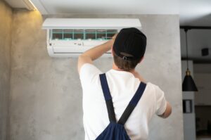 A man wearing overalls and a hat repairs an air conditioner, demonstrating skilled maintenance work.